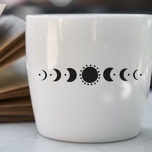 May include: White ceramic mug with a black sun and moon phases design. The design features a sun with a dotted circle around it and a series of crescent moons in different phases.