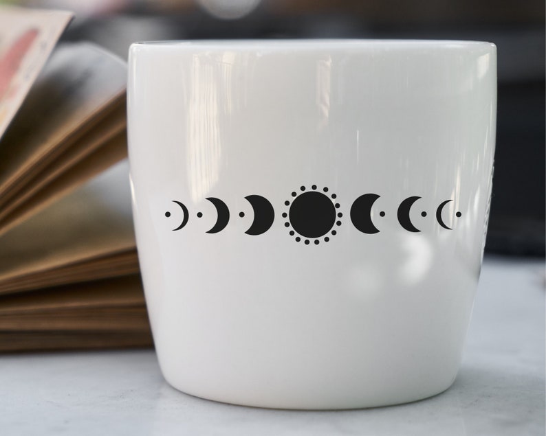 May include: White ceramic mug with a black sun and moon phases design. The design features a sun with a dotted circle around it and a series of crescent moons in different phases.