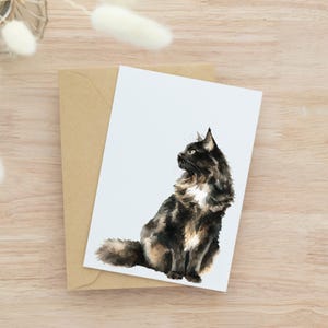 May include: Watercolor illustration of a fluffy black, brown, and white cat sitting on a white card. The card rests on a brown envelope, set against a light wood grain background. The cat is facing left, with its head turned.
