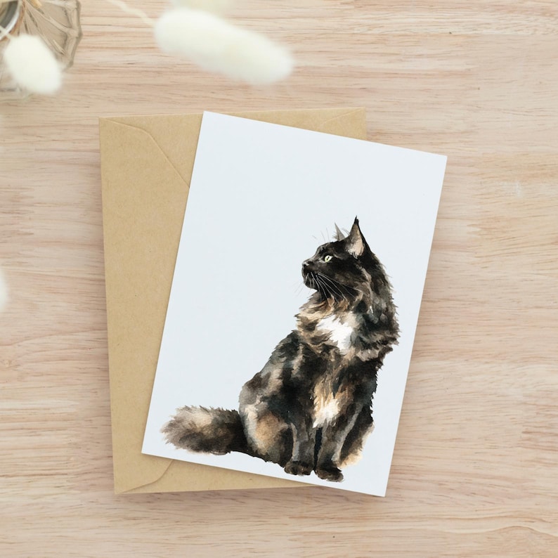 May include: Watercolor illustration of a fluffy black, brown, and white cat sitting on a white card. The card rests on a brown envelope, set against a light wood grain background. The cat is facing left, with its head turned.