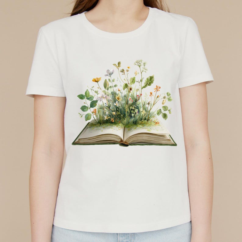 May include: White t-shirt featuring a watercolor illustration of an open book with colorful wildflowers and greenery emerging from the pages. The design includes yellow, orange, and purple flowers.