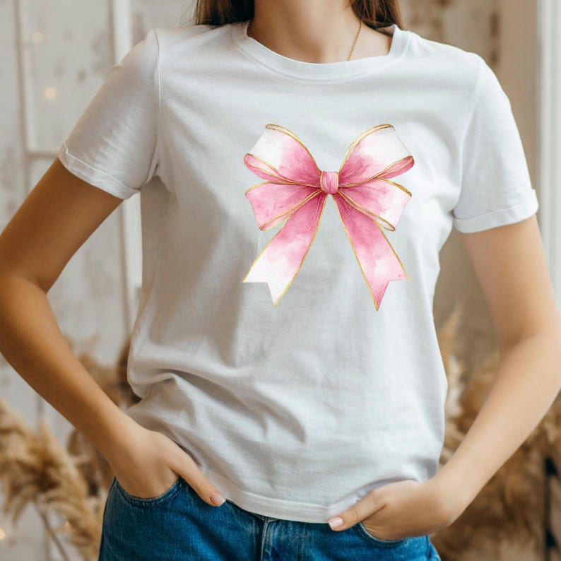 Puede incluir: Camiseta blanca con un lazo rosa estilo acuarela y ribetes dorados. El lazo est&aacute; centrado en el pecho. La camiseta tiene mangas cortas y cuello redondo.