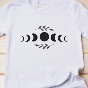 May include: White t-shirt with a black moon phases design. The design features a full moon, crescent moons, and a leafy branch.