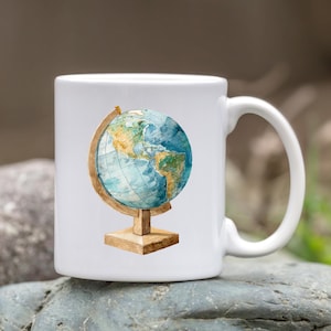 May include: White ceramic mug featuring a watercolor illustration of a globe. The globe shows the continents in shades of blue, green, and yellow, set on a wooden stand. The mug has a curved handle.