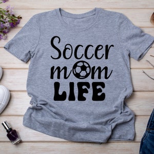 Op de afbeelding: Een lichtgrijs t-shirt met de tekst "Soccer mom LIFE" in zwarte letters en een voetbalafbeelding. Het shirt met korte mouwen is gemaakt van een zacht materiaal. Het shirt wordt gepresenteerd op een licht houten oppervlak.