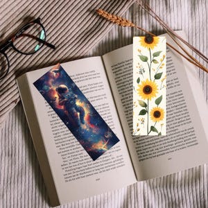 May include: Two bookmarks rest on an open book. One features an astronaut in space, the other has sunflowers. The astronaut bookmark has a colorful galaxy design. The sunflower bookmark has yellow flowers and green leaves.