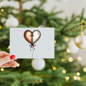 May include: A greeting card with a heart design, divided into brown and white halves, joined by a stitched seam. It has a decorative bow and string. The card is held in front of a Christmas tree with baubles.
