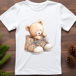 Puede incluir: Camiseta blanca con una ilustraci&oacute;n en acuarela de un oso de peluche dentro de un par de botas marrones. El oso es de color marr&oacute;n claro, con los ojos cerrados. Las botas tienen suelas y cordones blancos.