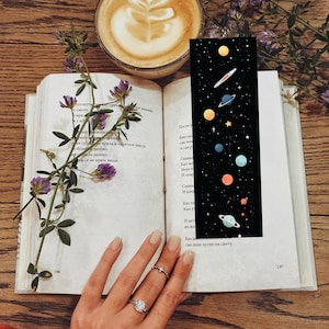 May include: A black bookmark with a colorful illustration of planets and stars. The bookmark is placed in an open book with text on the pages. A cup of coffee and purple flowers are also visible.