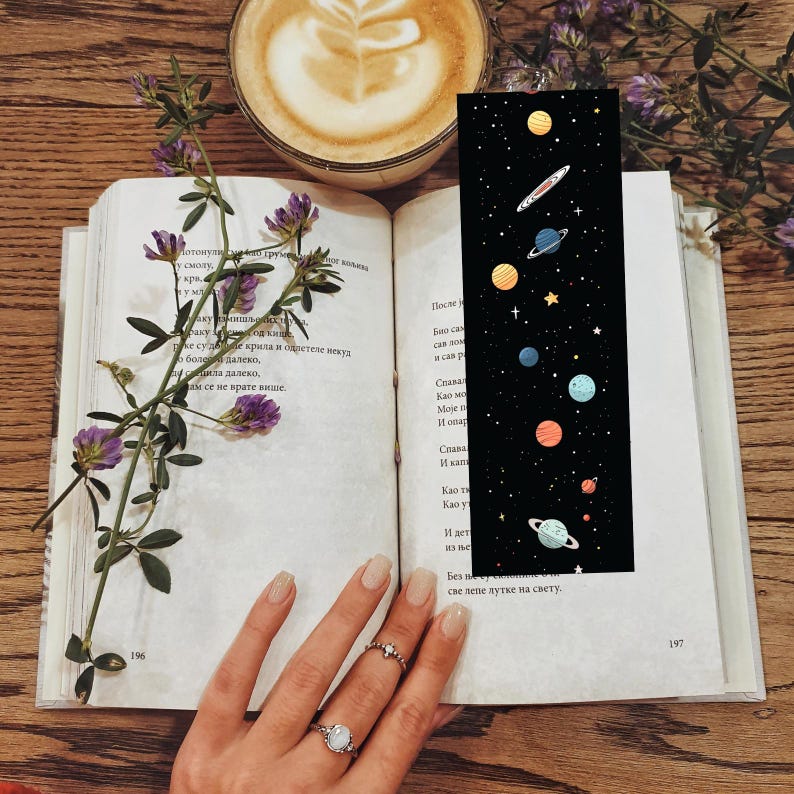 May include: A black bookmark with a colorful illustration of planets and stars. The bookmark is placed in an open book with text on the pages. A cup of coffee and purple flowers are also visible.
