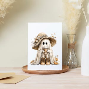 May include: A rectangular greeting card featuring a central illustration of a ghost wearing a wide-brimmed straw hat adorned with dried flowers. The ghost holds a tan coffee cup and wears tan lace-up boots, surrounded by small orange pumpkins.