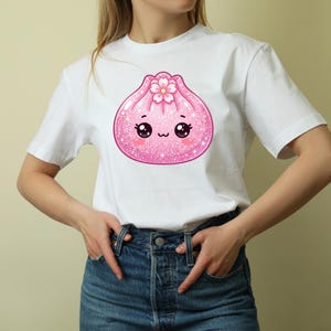 May include: A white short-sleeved t-shirt featuring a central graphic of a pink, glittery, cartoon-style dumpling character. The dumpling has large black eyes, a small mouth, rosy cheeks, and a pink cherry blossom flower on its top.
