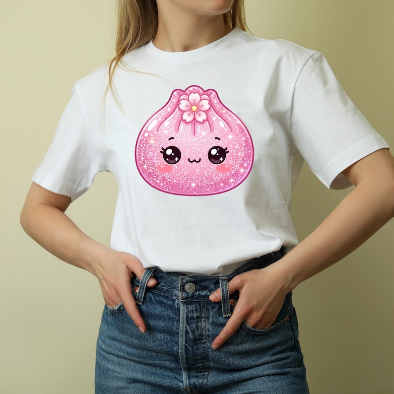 May include: A white short-sleeved t-shirt featuring a central graphic of a pink, glittery, cartoon-style dumpling character. The dumpling has large black eyes, a small mouth, rosy cheeks, and a pink cherry blossom flower on its top.