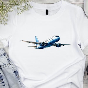 May include: White t-shirt featuring a watercolor illustration of a blue and white airplane in flight. The aircraft has detailed wings and engines, set against a clean white background. The design is centered on the shirt.