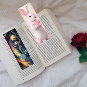 May include: Two bookmarks are placed in an open book. One bookmark features a white rabbit holding a pink heart. The other bookmark depicts an astronaut floating in space with a colorful galaxy background. A red rose is visible.