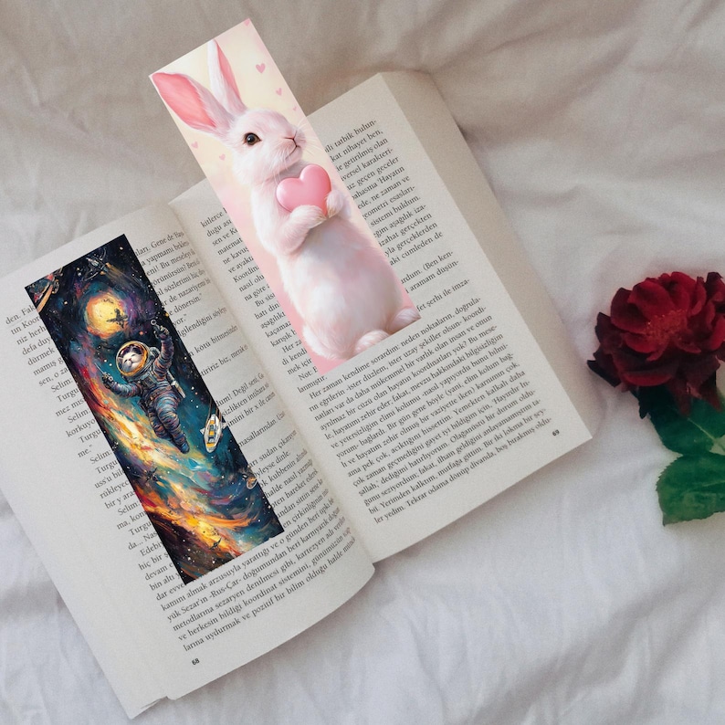 May include: Two bookmarks are placed in an open book. One bookmark features a white rabbit holding a pink heart. The other bookmark depicts an astronaut floating in space with a colorful galaxy background. A red rose is visible.