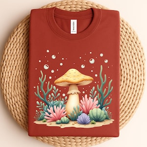 May include: Rust-colored crew neck sweatshirt featuring a whimsical mushroom graphic. The design includes a yellow mushroom with a cream-colored stem, surrounded by colorful coral and bubbles. The shirt is folded on a woven mat.