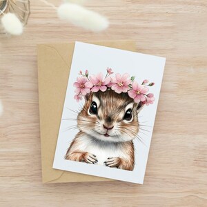 Op de afbeelding: Een wenskaart met een aquarel illustratie van een chipmunk met een roze bloemenkrans. De chipmunk heeft bruine vacht en grote zwarte ogen, tegen een lichtblauwe achtergrond. De kaart wordt geleverd met een bruine envelop.
