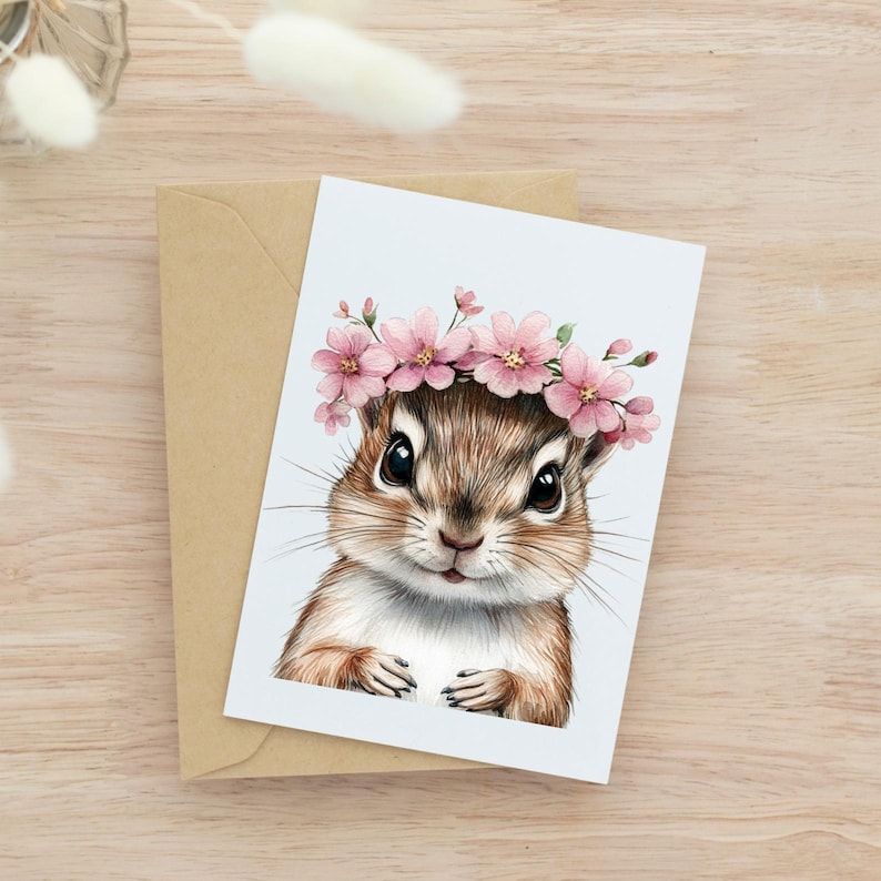 Op de afbeelding: Een wenskaart met een aquarel illustratie van een chipmunk met een roze bloemenkrans. De chipmunk heeft bruine vacht en grote zwarte ogen, tegen een lichtblauwe achtergrond. De kaart wordt geleverd met een bruine envelop.