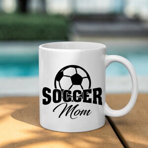 Op de afbeelding: Witte keramische mok met een zwart voetbalontwerp en de tekst "SOCCER Mom". De mok heeft een gebogen handvat en staat op een houten oppervlak. De achtergrond is wazig.