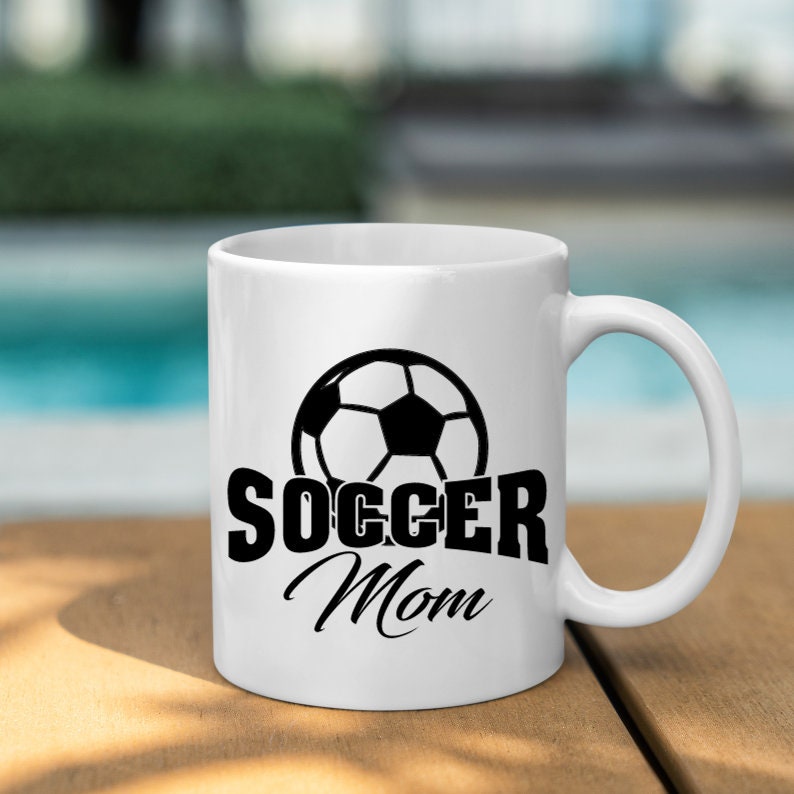 Op de afbeelding: Witte keramische mok met een zwart voetbalontwerp en de tekst "SOCCER Mom". De mok heeft een gebogen handvat en staat op een houten oppervlak. De achtergrond is wazig.