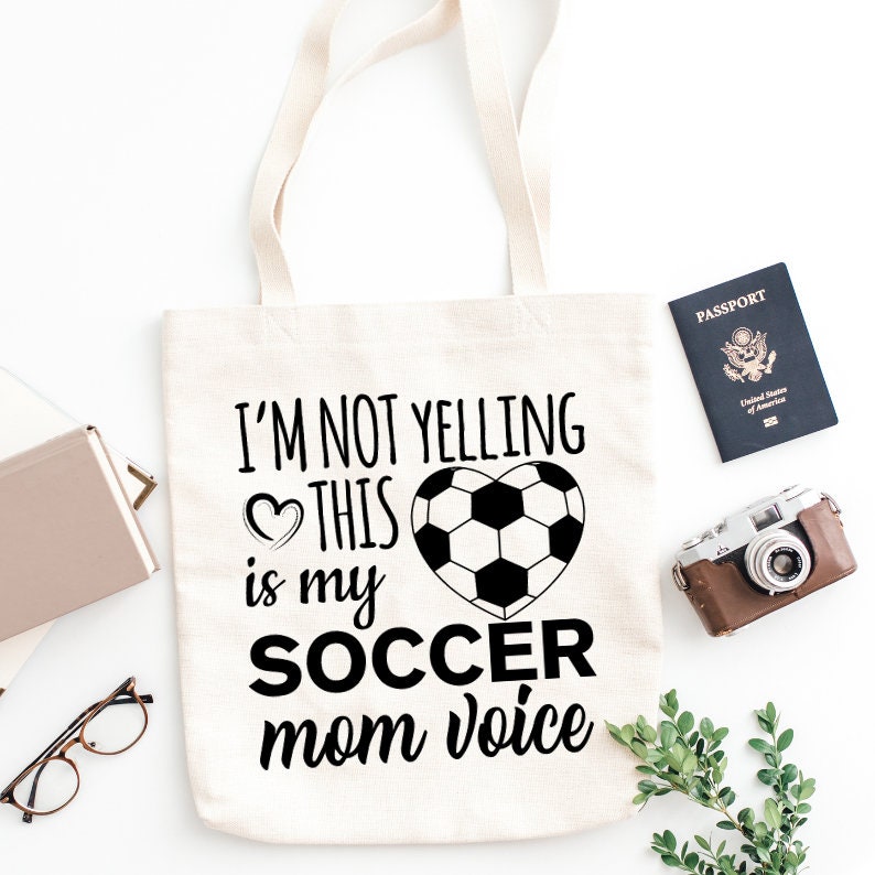 Op de afbeelding: Cr&egrave;mekleurige draagtas met de tekst "I'M NOT YELLING THIS is my SOCCER mom voice" en een voetbalhartontwerp. Een paspoort, camera, bril en een klein boek zijn ook zichtbaar.