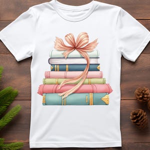 K&ouml;nnte beinhalten: Wei&szlig;es T-Shirt mit einem Aufdruck von einem Stapel bunter B&uuml;cher, die mit einer pfirsichfarbenen Schleife zusammengebunden sind. Die B&uuml;cher sind in Blau-, Rosa- und Gr&uuml;nt&ouml;nen gehalten und haben goldene Akzente. Ein verspieltes Design f&uuml;r B&uuml;cherliebhaber.