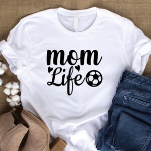 Op de afbeelding: Wit T-shirt met de zwarte tekst "mom Life" en een voetbalafbeelding. Het shirt wordt gecombineerd met een blauwe jeans en bruine laarzen, wat een casual stijl suggereert. Het ontwerp is eenvoudig en opvallend.