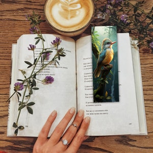 May include: A close-up of a book with a bookmark featuring a blue and yellow bird perched on a branch. The bookmark is set against a background of green foliage. The book is open to a page with text in a foreign language.