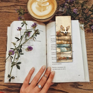 May include: A bookmark with a watercolour illustration of a Corgi dog looking over a pile of books. The bookmark is in an open book with text, beside a coffee cup and purple flowers. The scene is set on a wooden surface.