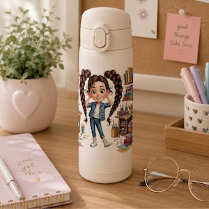 May include: A cream-coloured insulated water bottle featuring a graphic illustration of a person with long brown pigtails wearing a denim jacket and jeans, standing in an art studio filled with various painting supplies and colourful art materials.