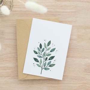 May include: A white greeting card with a watercolor illustration of a green leafy branch, resting on a brown envelope. The card features various shades of green leaves and small gray dots. The background is a light brown wooden surface.