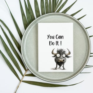 May include: A white card with the text "You Can Do It!" printed in black. A cartoon illustration of a black bull with large eyes is below the text.