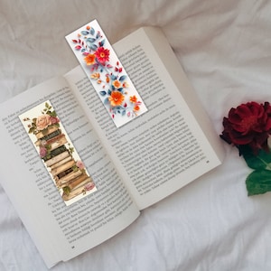 May include: Two decorative bookmarks with floral designs are placed on an open book. One bookmark shows a stack of books with roses, and the other has orange flowers and blue leaves. A red rose is in the background.