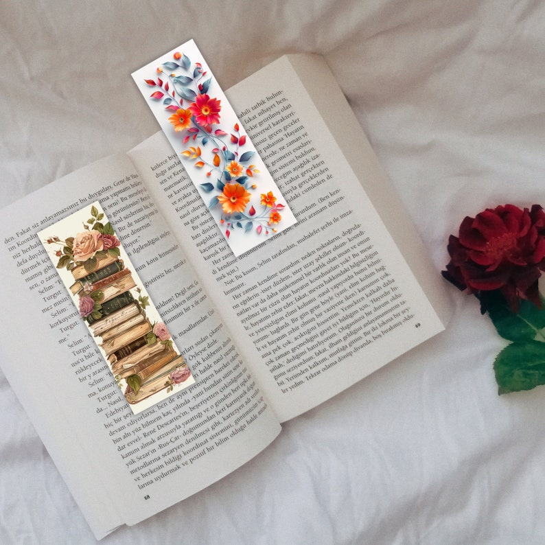 May include: Two decorative bookmarks with floral designs are placed on an open book. One bookmark shows a stack of books with roses, and the other has orange flowers and blue leaves. A red rose is in the background.