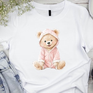 Puede incluir: Camiseta blanca con una ilustraci&oacute;n en acuarela de un oso de peluche con un mono rosa con capucha. El oso est&aacute; sentado con las piernas cruzadas y sonriendo. El dise&ntilde;o est&aacute; centrado en la camiseta.