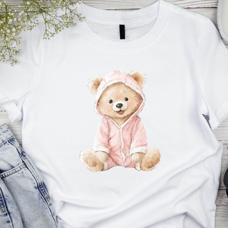 Puede incluir: Camiseta blanca con una ilustraci&oacute;n en acuarela de un oso de peluche con un mono rosa con capucha. El oso est&aacute; sentado con las piernas cruzadas y sonriendo. El dise&ntilde;o est&aacute; centrado en la camiseta.