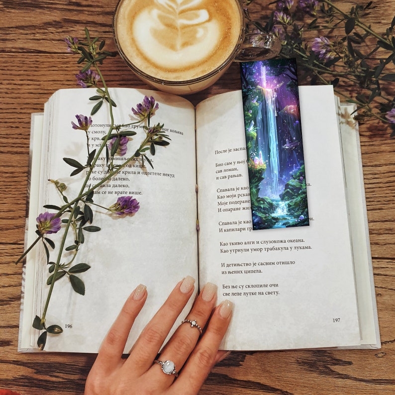 May include: A close-up of an open book with a bookmark featuring a colorful waterfall scene. The book is resting on a wooden table with a cup of coffee and a bouquet of purple flowers.