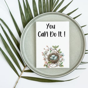 May include: A white card with the text "You Can Do It!" in black lettering. The card features a watercolor illustration of a bird's nest with three blue eggs surrounded by white flowers.