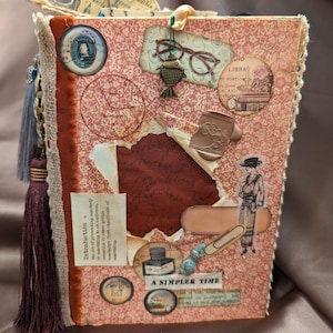 May include: A vintage-style journal with a brown leather cover and a pink patterned paper overlay. The journal is decorated with various embellishments, including a small metal fish, a pair of glasses, a paper tag with the word "Libra", and a small paper tag with the text "A Simpler Time".