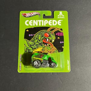 May include: A Hot Wheels Cool-One die-cast vehicle in lime green packaging featuring Atari Centipede branding. The card displays the Centipede game artwork, while the toy car features matching green paint, chrome engine details, and Atari logos.