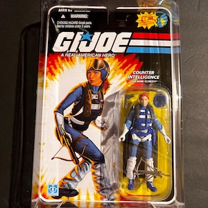 May include: A G.I. Joe action figure of Scarlett, a counter-intelligence operative, in a clear plastic package. The figure is wearing a blue and white uniform with a crossbow. The packaging features the G.I. Joe logo and the text "A Real American Hero."