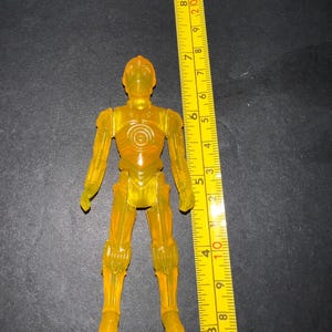 May include: A translucent yellow C-3PO action figure, approximately 5 inches tall, stands upright. The figure has detailed features and is positioned next to a yellow ruler with black markings. The background is a dark surface.