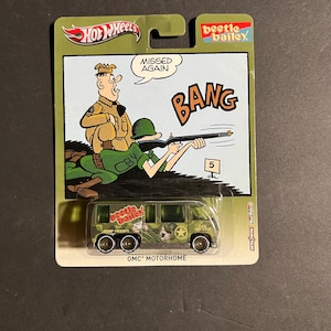 Könnte beinhalten: Ein Hot Wheels GMC Motorhome Spielzeugfahrzeug in olivgrüner Verpackung mit Beetle Bailey Comic-Grafik. Die Karte zeigt einen Soldaten, der mit einem Gewehr schießt, mit dem Text MISSED AGAIN und BANG. Der Van trägt Beetle Bailey Branding und Camp Swampy.