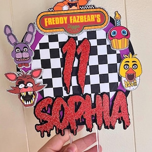May include: A custom birthday cake topper featuring characters from Freddy Fazbear's Pizza. The topper includes the text "Freddy Fazbear's Pizza Place", the name "Sophia", and the number "11" in red glitter. Additional character cutouts include a cupcake, a chicken, a rabbit, and a fox.