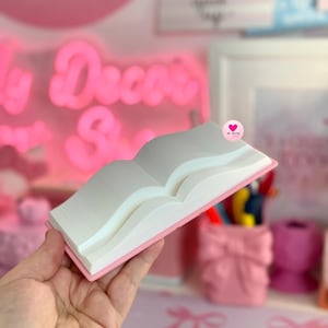 May include: A pink and white decorative book, open to display the pages. The book has a pink border and white pages. The background includes a pink neon sign that says "My Decor Shop".