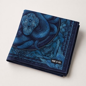 May include: Folded square cloth featuring a detailed illustration of a blue serpent. The artwork has intricate patterns and shading, with the text "VIBE & CO." on a small black label. The color palette is primarily shades of blue.