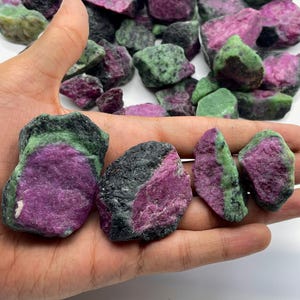 Peut inclure: Une collection de pierres brutes de rubis zoisite. Les pierres présentent un mélange de couleurs, notamment le magenta profond, le vert forêt et le noir. Les pierres sont tenues dans une main, avec d'autres pierres dispersées en arrière-plan.