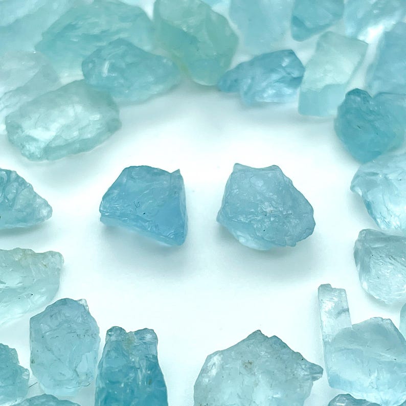 Op de afbeelding: Een verzameling ruwe, aquamarijnkleurige edelstenen. De stenen vari&euml;ren in grootte en vorm, met een doorschijnende kwaliteit. De edelstenen, in verschillende blauwtinten, zijn gerangschikt op een witte ondergrond, wat een natuurlijke uitstraling cre&euml;ert.