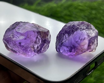 May include: Two raw, unpolished amethyst crystals with a deep purple hue and translucent, jagged surfaces. These natural gemstones feature irregular, multifaceted shapes with visible internal fractures and varying shades of violet throughout.
